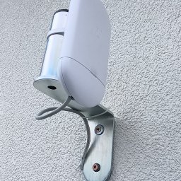 Bogdan Popłoński Clever-Tech - Biały czujnik ruchu, zamontowany na metalowym uchwycie na teksturowanej, jasnej ścianie zewnętrznej budynku. Widoczne śruby mocujące i szary kabel.