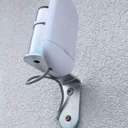 Biały czujnik ruchu, zamontowany na metalowym uchwycie na teksturowanej, jasnej ścianie zewnętrznej budynku. Widoczne śruby mocujące i szary kabel.