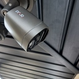 Bogdan Popłoński Clever-Tech - Szara kamera monitoringu Pixir zamontowana pod ciemnym podbitką dachową z widocznymi śrubami mocującymi. Widok kamery pod kątem, w tle fragment ściany budynku.