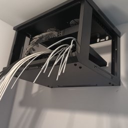 Bogdan Popłoński Clever-Tech - Czarna szafa rack wisząca na ścianie z kablami sieciowymi, routerem i wentylatorami. Widok od dołu, instalacja sieci komputerowej w biurze lub domu.
