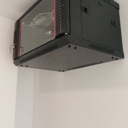 Bogdan Popłoński Clever-Tech - Czarna szafa rack wisząca na białej ścianie, z widocznymi kablami wewnątrz. Zamknięta na klucz, z perforowanymi bokami i szklanymi drzwiami.