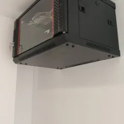 Czarna szafa rack wisząca na białej ścianie, z widocznymi kablami wewnątrz. Zamknięta na klucz, z perforowanymi bokami i szklanymi drzwiami.