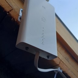 Bogdan Popłoński Clever-Tech - Zewnętrzna antena 5G ZTE zamontowana na drewnianej elewacji budynku, z widocznymi kablami sygnałowymi i zasilającymi. Widok z dołu, błękitne niebo w tle.
