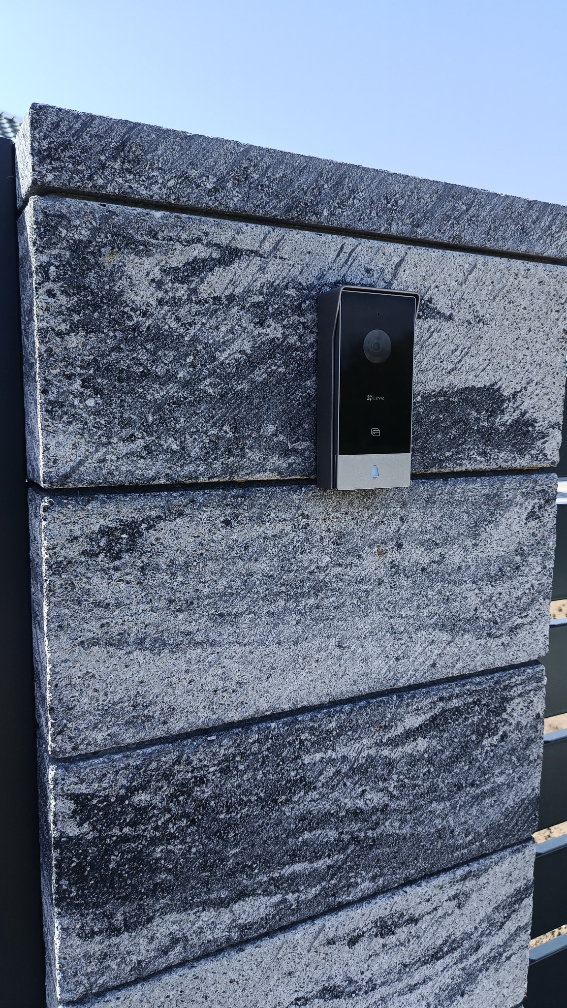 Czarny wideodomofon Ezviz zamontowany na szarym, kamiennym słupku ogrodzeniowym. Widoczny obiektyw kamery i przycisk dzwonka. Słoneczny dzień.