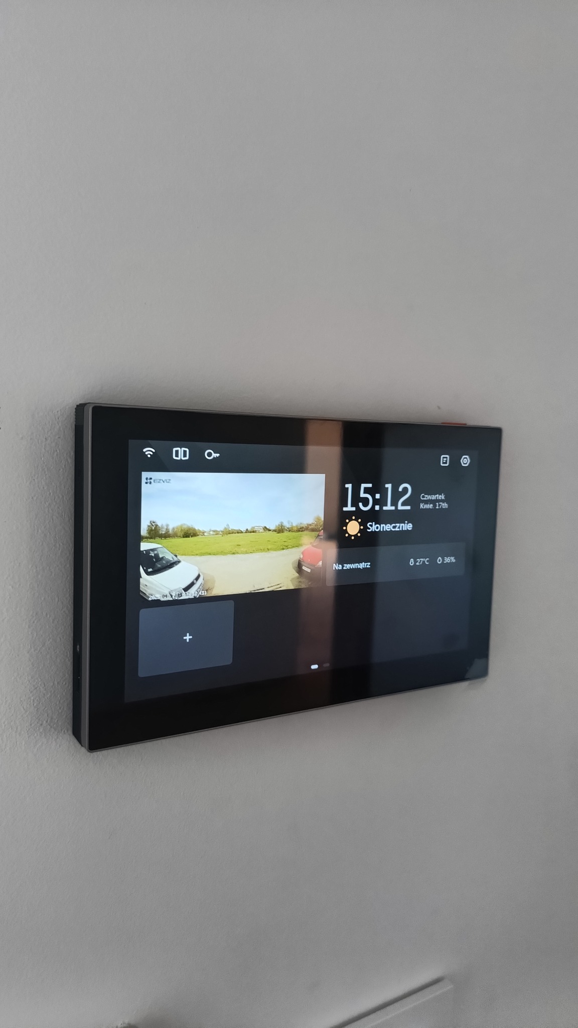 Panel dotykowy systemu monitoringu na ścianie, prezentujący obraz z kamery na zewnątrz, z informacjami o pogodzie i dacie. Widoczne auta na podjeździe.