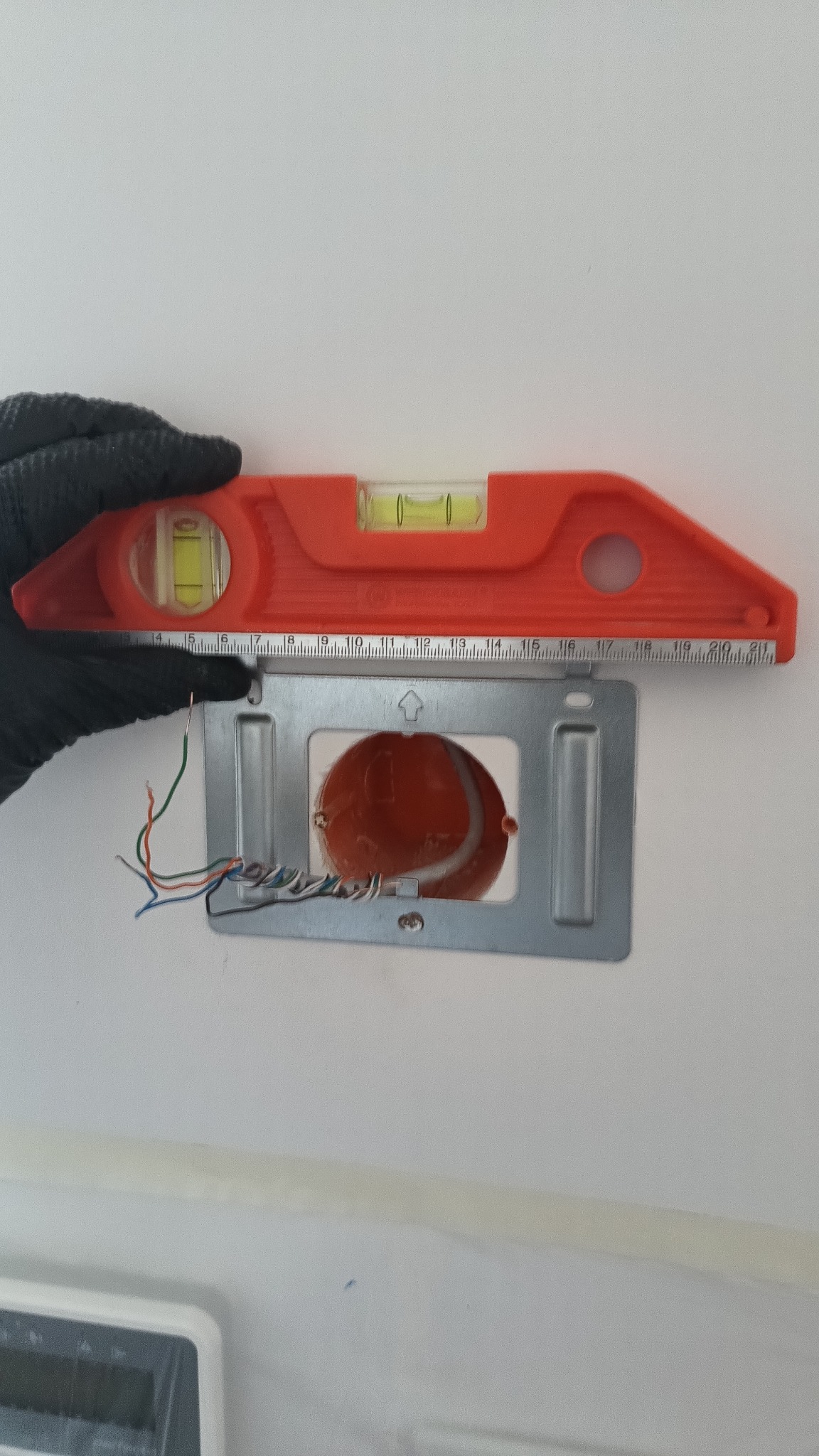 Instalacja puszki elektrycznej w ścianie, poziomowana pomarańczową poziomicą. Widoczne przewody i ręka w czarnej rękawicy.