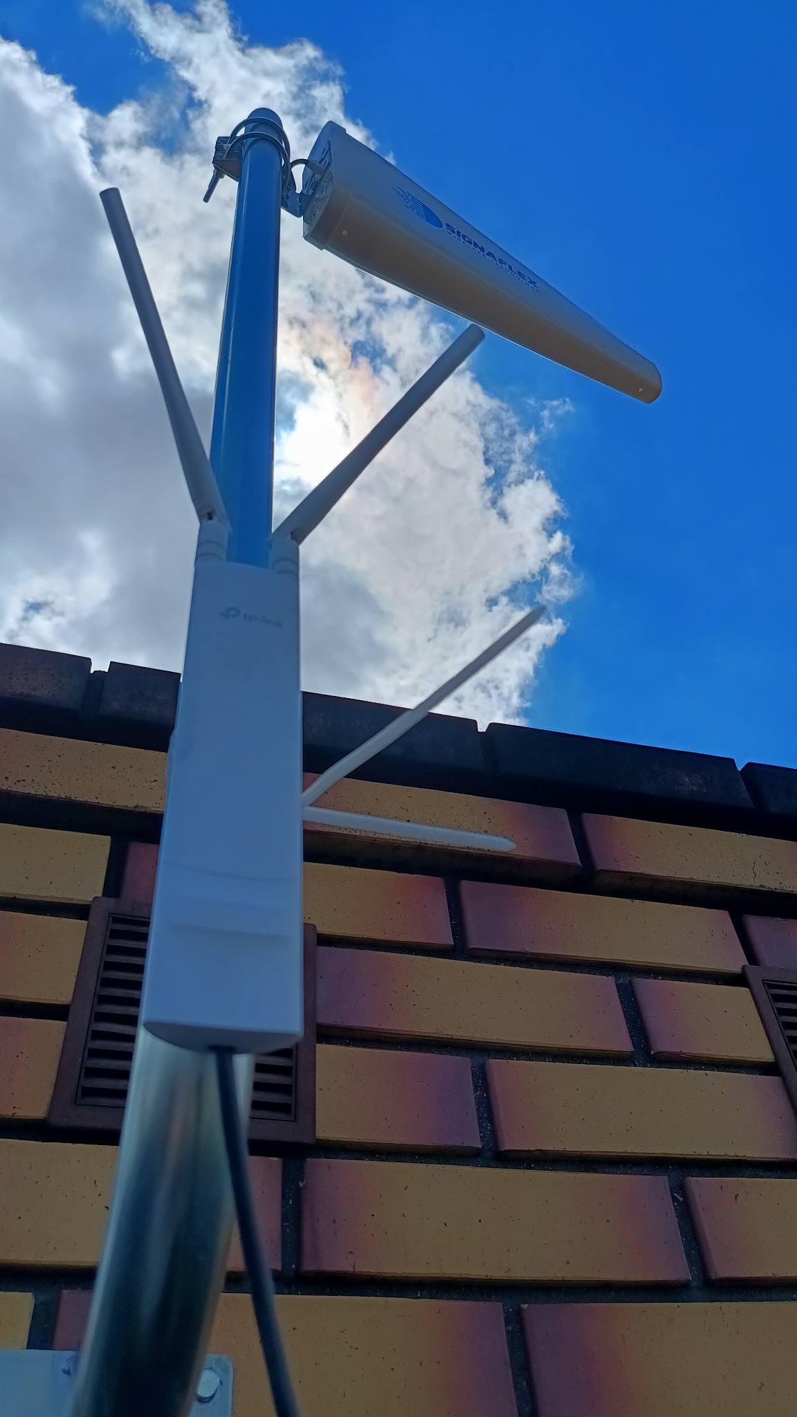 Antena telekomunikacyjna Signaflex na tle błękitnego nieba, zamontowana na budynku w Białej Rawskiej. Widok z dołu, skierowany ku górze.