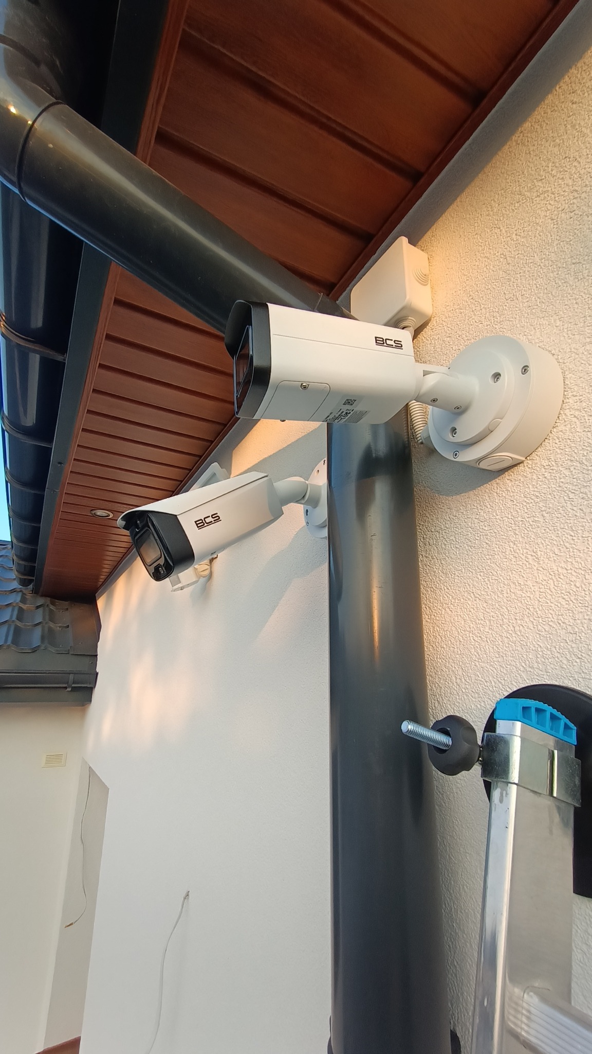 Dwie kamery BCS zamontowane na elewacji budynku, z widoczną rurą spustową i fragmentem drabiny. Oświetlenie popołudniowe, ciepłe barwy.