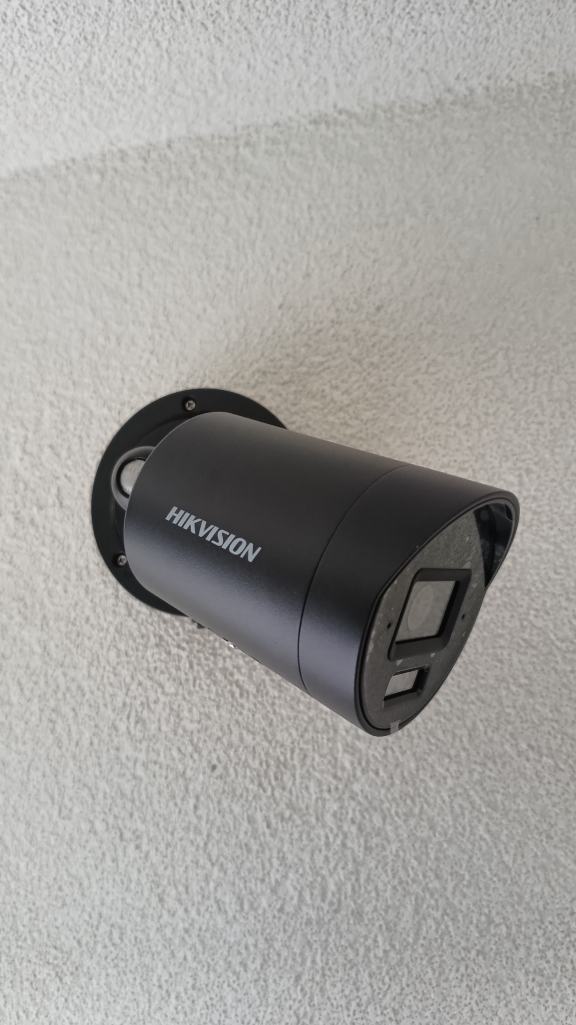 Czarna kamera Hikvision zamontowana na teksturowanej, jasnej ścianie. Widoczny obiektyw kamery i logo producenta. Kąt ujęcia lekko z dołu.
