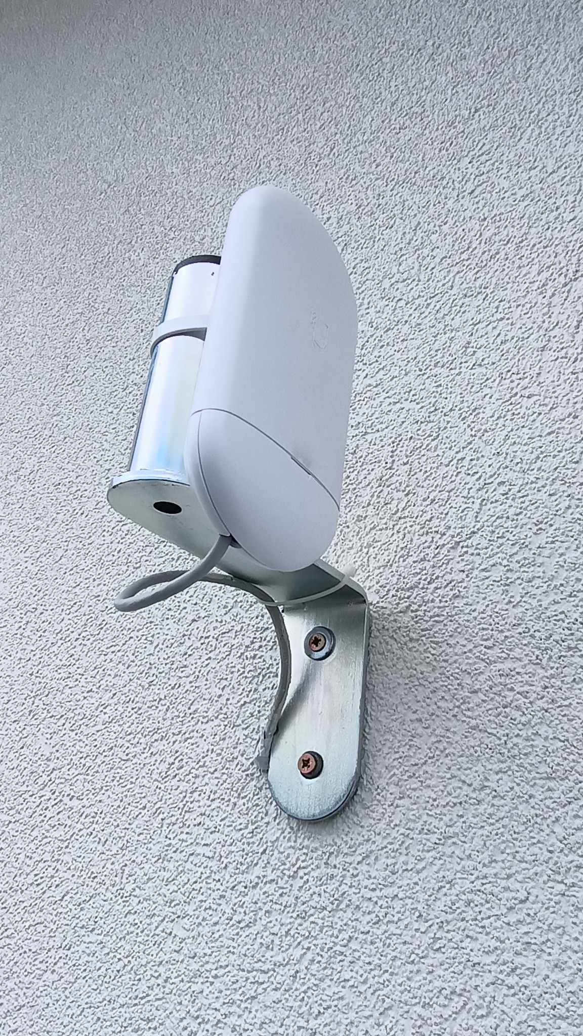 Biały czujnik ruchu, zamontowany na metalowym uchwycie na teksturowanej, jasnej ścianie zewnętrznej budynku. Widoczne śruby mocujące i szary kabel.