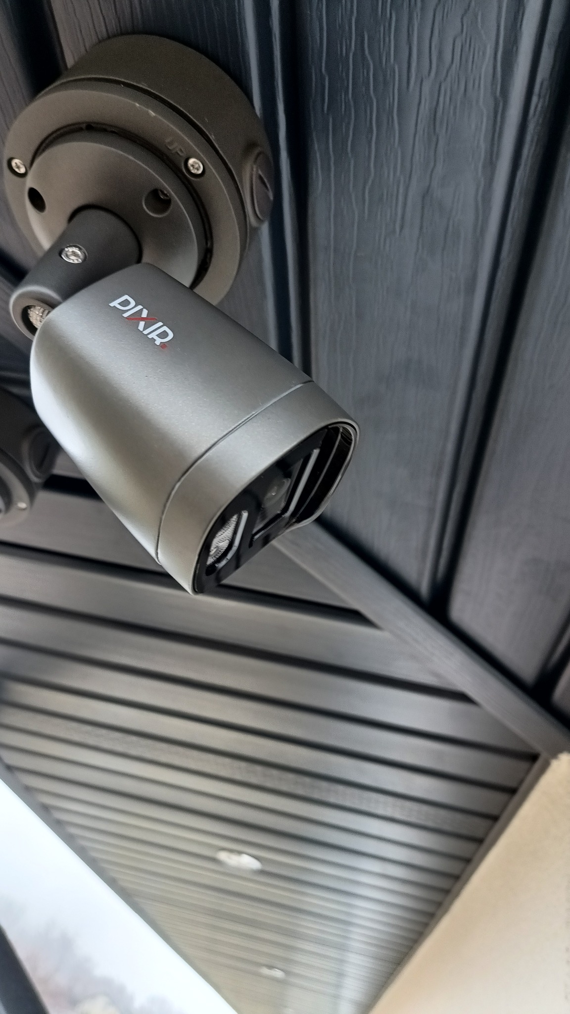 Szara kamera monitoringu Pixir zamontowana pod ciemnym podbitką dachową z widocznymi śrubami mocującymi. Widok kamery pod kątem, w tle fragment ściany budynku.