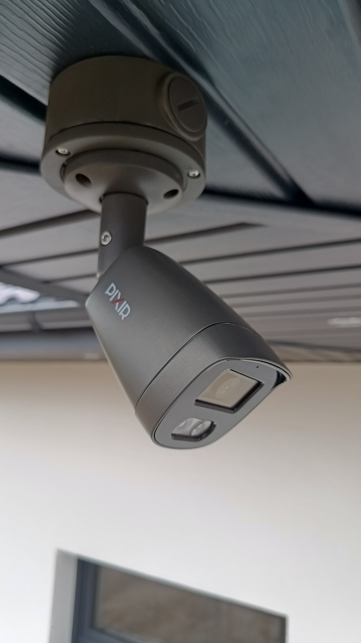 Szara kamera monitoringu PixIR zamontowana pod drewnianym sufitem, skierowana w dół. Widoczny obiektyw i diody IR. Ujęcie z dołu, widoczne mocowanie kamery.