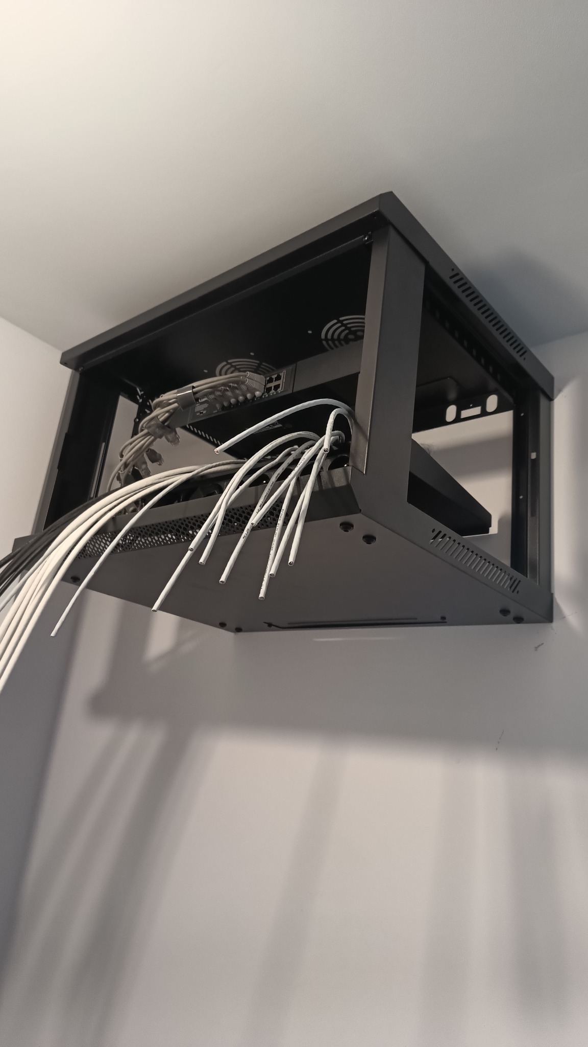 Czarna szafa rack wisząca na ścianie z kablami sieciowymi, routerem i wentylatorami. Widok od dołu, instalacja sieci komputerowej w biurze lub domu.
