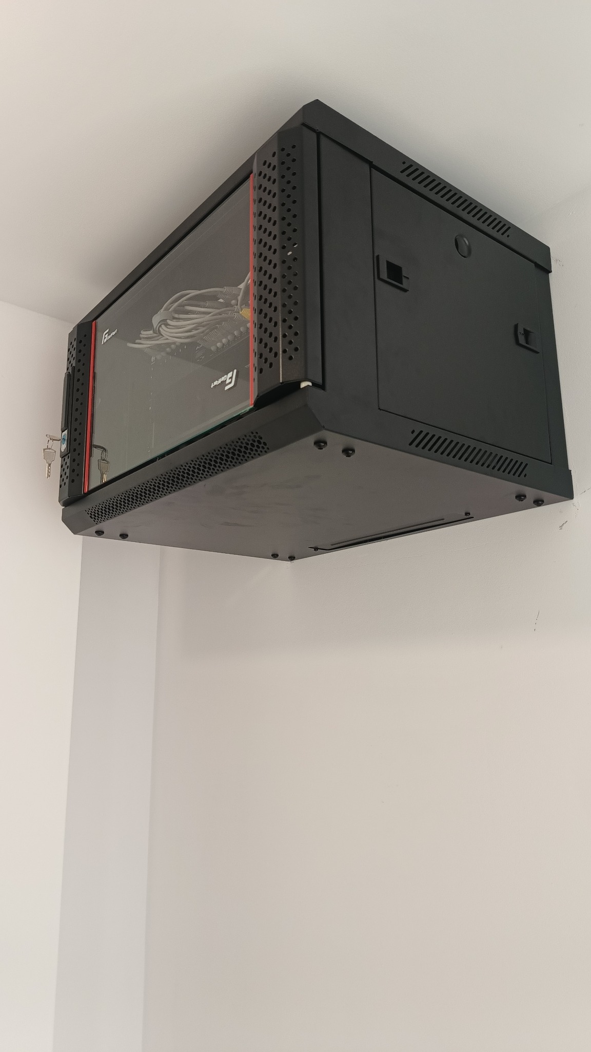 Czarna szafa rack wisząca na białej ścianie, z widocznymi kablami wewnątrz. Zamknięta na klucz, z perforowanymi bokami i szklanymi drzwiami.