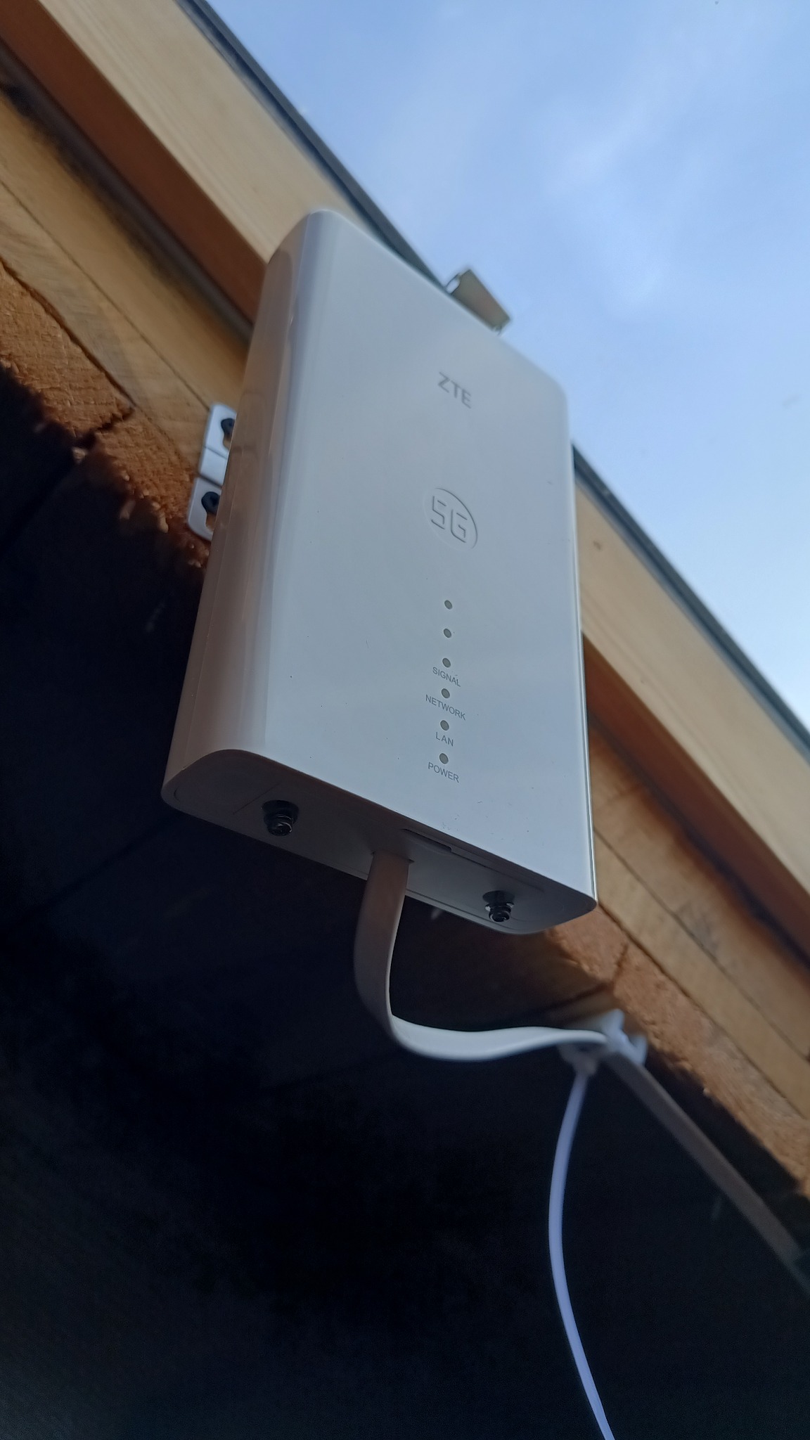 Zewnętrzna antena 5G ZTE zamontowana na drewnianej elewacji budynku, z widocznymi kablami sygnałowymi i zasilającymi. Widok z dołu, błękitne niebo w tle.
