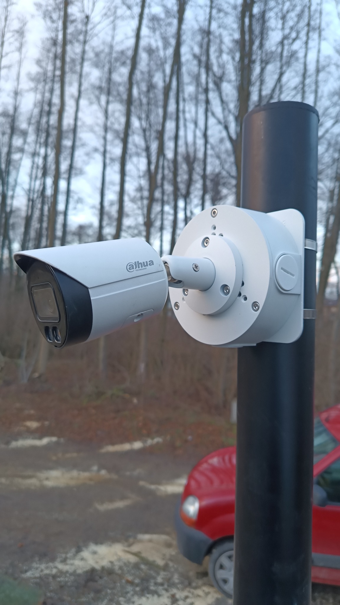 Kamera monitoringu Dahua zamontowana na czarnym słupku, widoczna na tle drzew i czerwonego samochodu. Obudowa kamery w kolorze jasnoszarym.