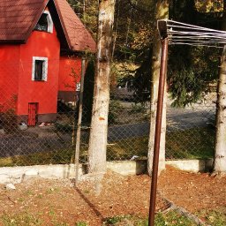 LPO-PK Las Park Ogród Usługi Ogrodnicze - Fragment posesji z czerwonym domem, drzewami i metalowym słupkiem na tle siatki ogrodzeniowej, jesienne liście na trawie.