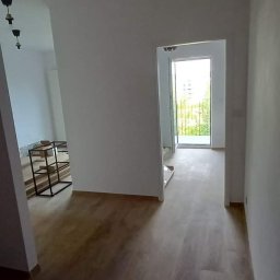 Patryk Sandecki - Wnętrze po remoncie: świeżo pomalowane białe ściany, nowa drewniana podłoga, widoczne niedokończone prace w tle i surowe lampy bez kloszy.