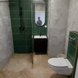 Patryk Sandecki - Wykończona łazienka z zielonymi płytkami, kabiną prysznicową typu walk-in z czarną armaturą, wiszącą toaletą i wzorzystą podłogą z płytek imitujących drewno, odbicie fotografa w owalnym lustrze.