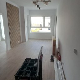 Patryk Sandecki - Wnętrze w trakcie prac wykończeniowych: widoczna drewniana listwa dekoracyjna na ścianie, lampa wisząca z okrągłymi kloszami, podłoga wyłożona panelami oraz skrzynki narzędziowe na podłodze.