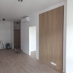 Patryk Sandecki - Jasne panele podłogowe w pustym pokoju z drewnianą ścianką działową z listewek i czarnymi reflektorami sufitowymi