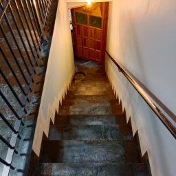 Patryk Sandecki - Schody wyłożone szarymi płytkami imitującymi kamień, z metalową balustradą po lewej i drewnianą poręczą po prawej stronie, prowadzące do uchylonych, drewnianych drzwi z szybkami.