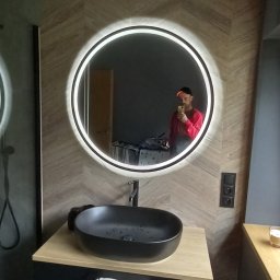 Patryk Sandecki - Nowoczesna łazienka z okrągłym lustrem podświetlanym LED, czarną umywalką nablatową i drewnopodobną okładziną ścienną ułożoną we wzór jodełki. W lustrze odbija się osoba robiąca zdjęcie.