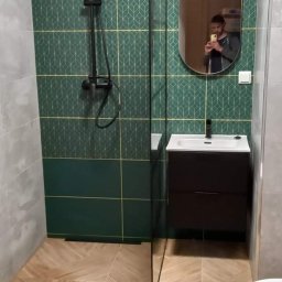 Patryk Sandecki - Nowoczesna łazienka z zielonymi płytkami w geometryczny wzór, czarną armaturą i drewnopodobną podłogą ułożoną w jodełkę, widok na kabinę prysznicową i umywalkę z szafką, odbicie fotografa w owalnym...