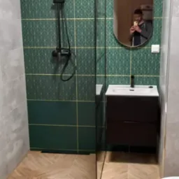 Nowoczesna łazienka z zielonymi płytkami w geometryczny wzór, czarną armaturą i drewnopodobną podłogą ułożoną w jodełkę, widok na kabinę prysznicową i umywalkę z szafką, odbicie fotografa w owalnym...