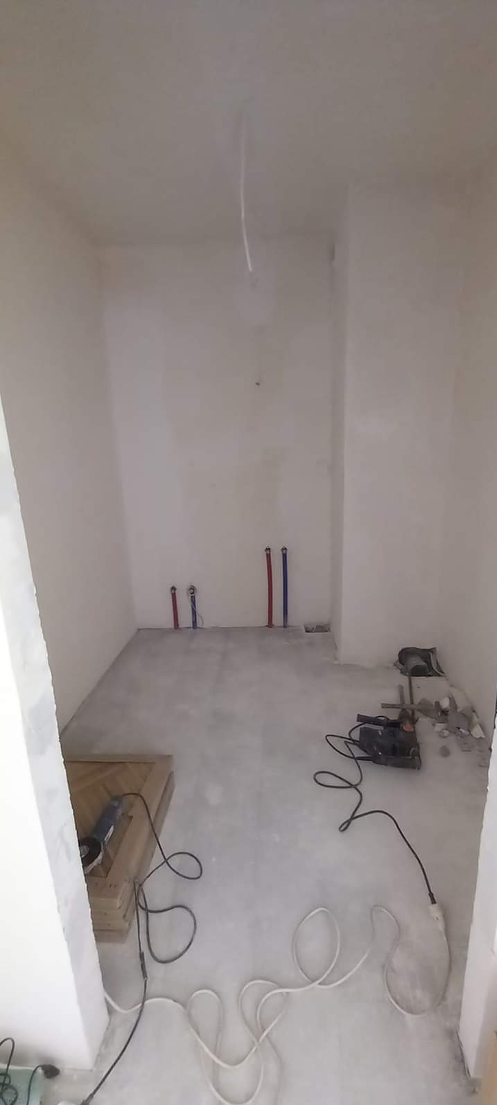 Surowe wnętrze łazienki w trakcie prac: widoczne piony instalacji wodnej, szpachlowane ściany, wylewka na podłodze i porozrzucane narzędzia budowlane.