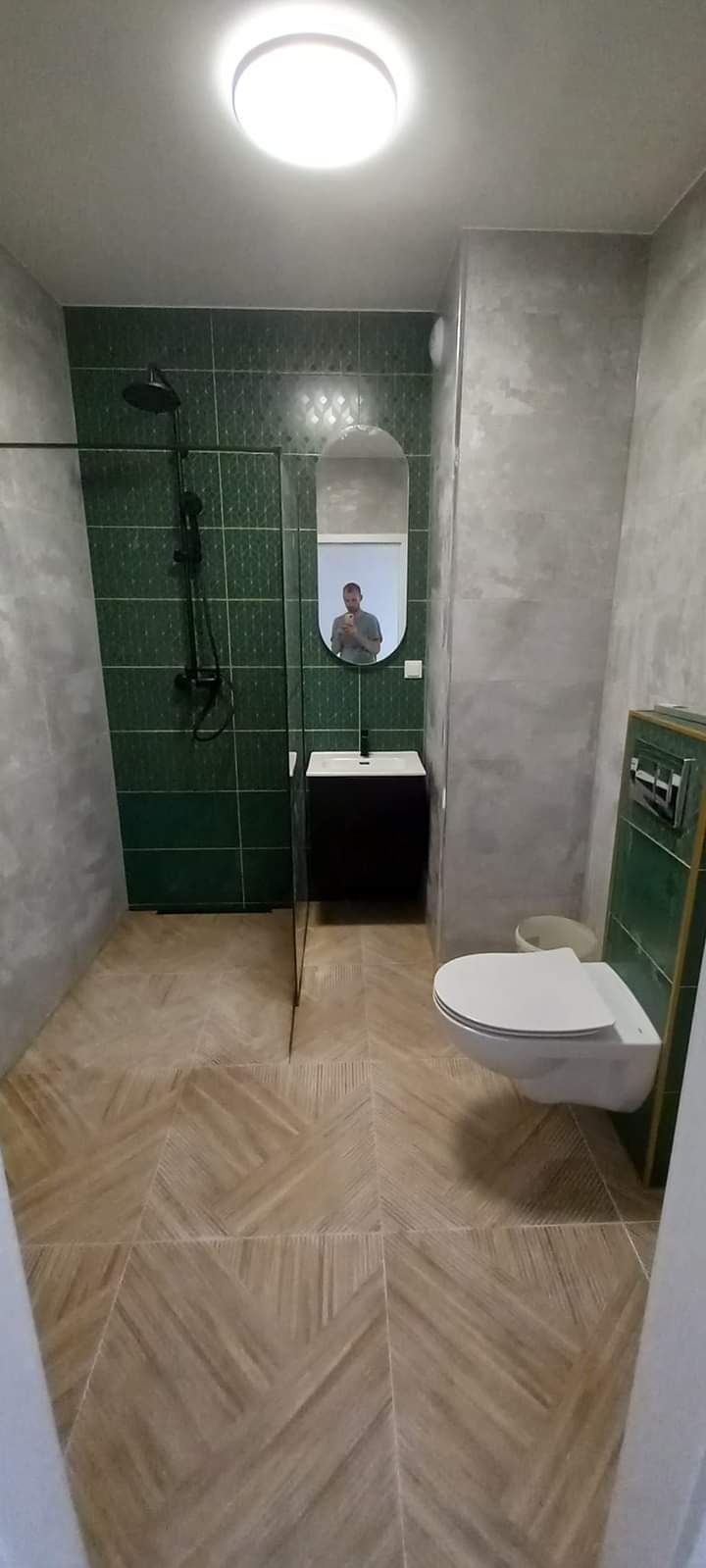 Wykończona łazienka z zielonymi płytkami, kabiną prysznicową typu walk-in z czarną armaturą, wiszącą toaletą i wzorzystą podłogą z płytek imitujących drewno, odbicie fotografa w owalnym lustrze.