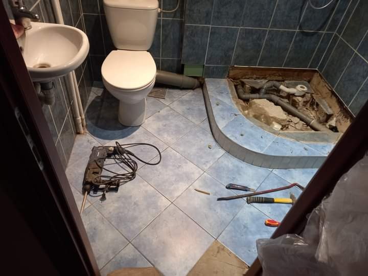 Niefortunny kadr z remontowanej łazienki: widok na zdemontowany brodzik z odsłoniętą instalacją hydrauliczną, obok leżą narzędzia, a w tle umywalka i toaleta na niebieskich płytkach.