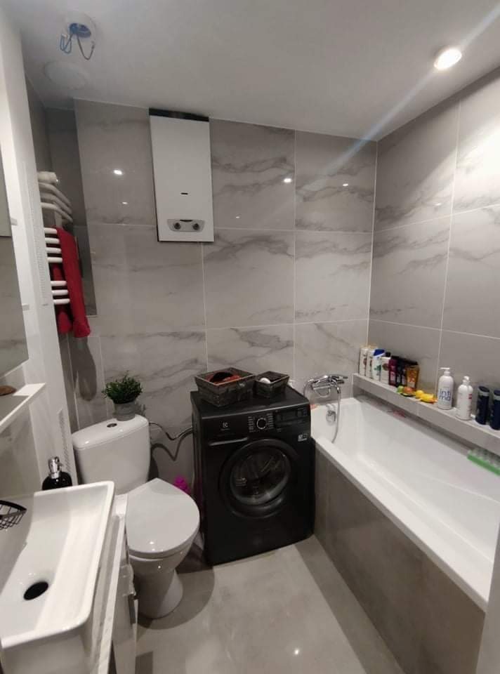 Ukończona łazienka w szaro-białej tonacji z pralką w kolorze czarnym, umywalką na szafce, toaletą i wanną z półką na kosmetyki.