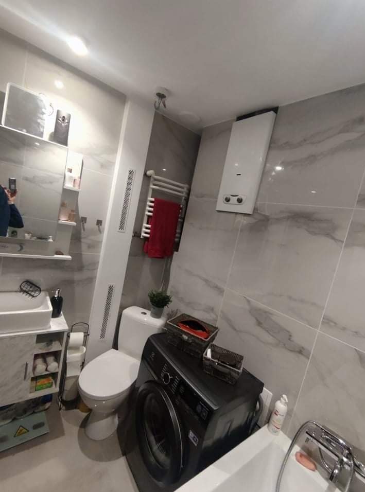 Nowoczesna łazienka w szarościach z marmurkowymi płytkami, widoczna umywalka z szafką, toaleta, grzejnik drabinkowy z czerwonym ręcznikiem, piec gazowy oraz pralka z koszykami na górze.