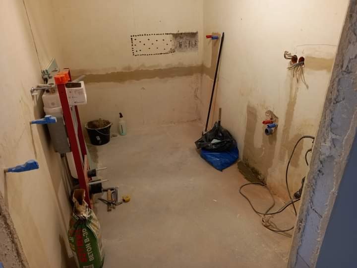 Łazienka w trakcie remontu: odsłonięte instalacje wodne i elektryczne, ściany przygotowane do dalszych prac, widoczny worek z betonem B-30, drabina i inne narzędzia.