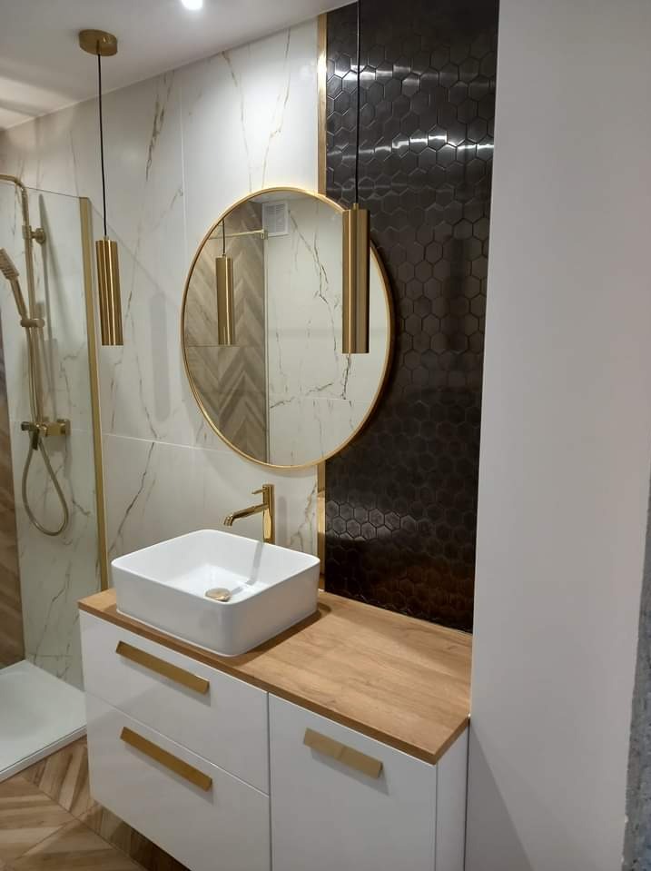 Nowoczesna łazienka z okrągłym lustrem w złotej ramie, wiszącymi złotymi lampami, białą kwadratową umywalką nablatową na szafce z drewnianym blatem i złotymi uchwytami, oraz czarną ścianą...