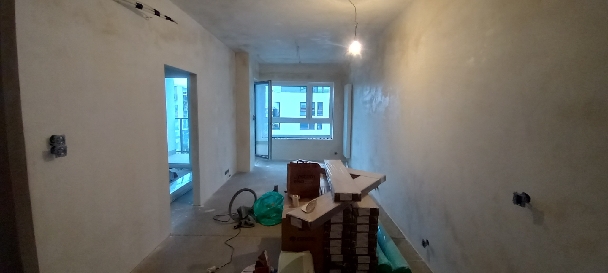 Wnętrze mieszkania w trakcie prac wykończeniowych; ściany po szpachlowaniu, widoczne puszki elektryczne, podłoga przygotowana pod panele, materiały budowlane w tle, okno balkonowe otwarte.
