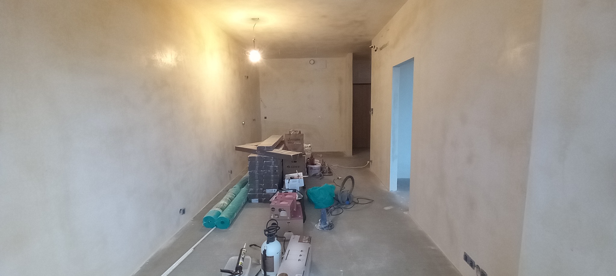 Wnętrze w trakcie prac wykończeniowych: ściany pokryte szpachlą o nieregularnej fakturze, widoczne narzędzia i materiały budowlane, surowe oświetlenie, przygotowane do dalszych etapów.