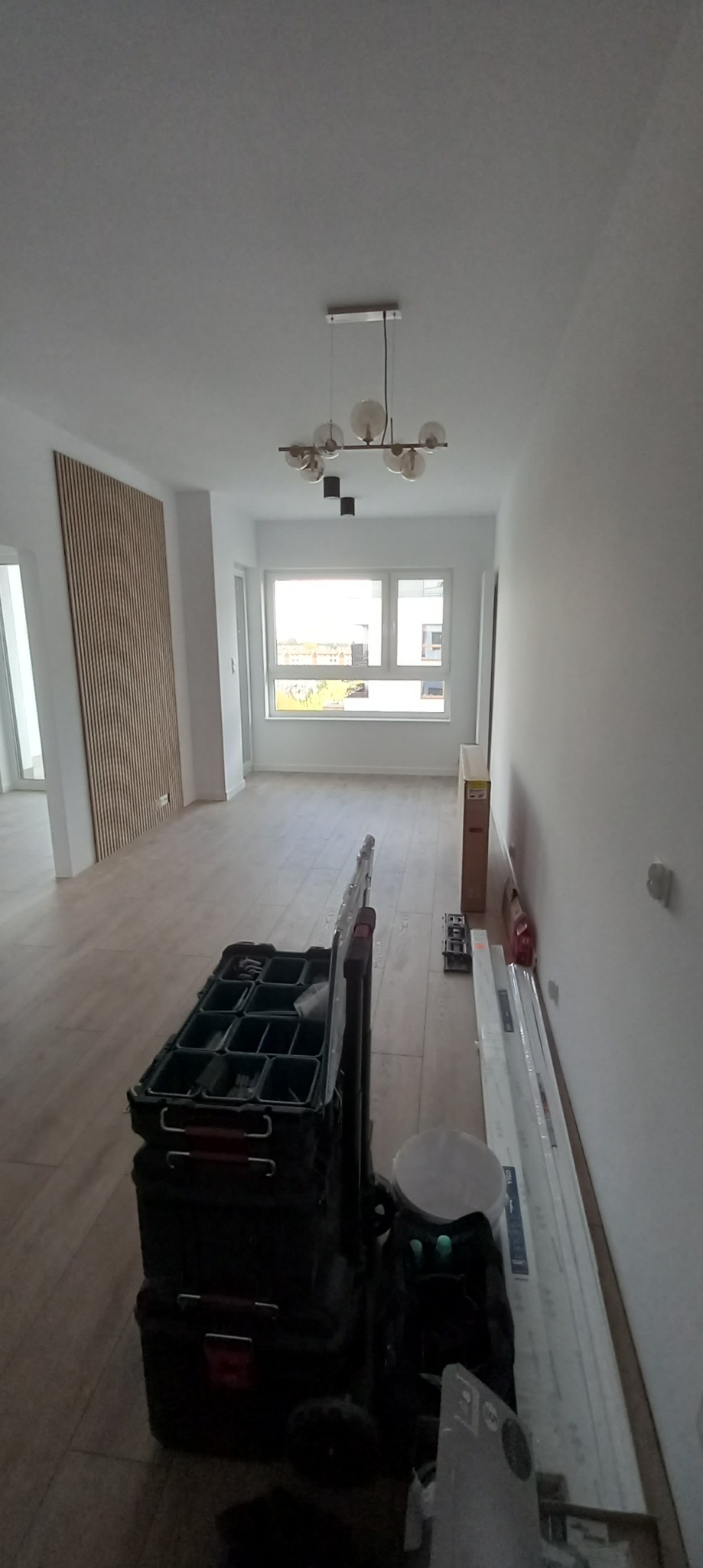 Wnętrze w trakcie prac wykończeniowych: widoczna drewniana listwa dekoracyjna na ścianie, lampa wisząca z okrągłymi kloszami, podłoga wyłożona panelami oraz skrzynki narzędziowe na podłodze.