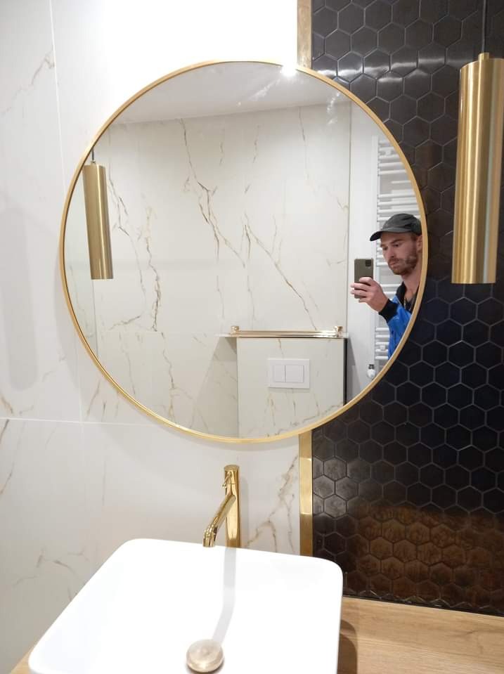 Nowoczesna łazienka z okrągłym lustrem w złotej ramie, białą umywalką i baterią w kolorze złota, biało-złotymi płytkami imitującymi marmur oraz czarnymi heksagonalnymi płytkami na ścianie,...