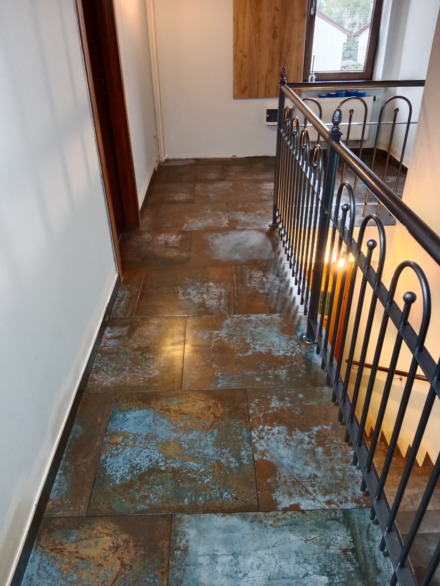 Korytarz z podłogą wyłożoną płytkami imitującymi rdzę, z czarną metalową balustradą schodów i widokiem na okno.