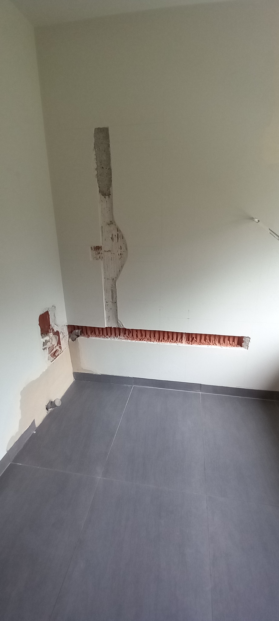 Przygotowanie bruzd w ścianie pod instalację hydrauliczną w łazience, widoczne szare płytki podłogowe i fragment rury wystającej ze ściany.