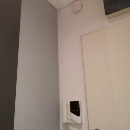 Jacek Osuch - Fragment wnętrza po odświeżeniu: Szara ściana z ciemniejszym pasem, biała listwa sufitowa, domofon przy białych drzwiach z metalową klamką i kluczami.