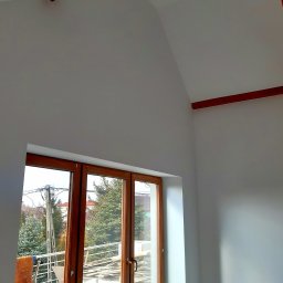 Jacek Osuch - Pomalowane na biało poddasze z drewnianymi belkami na suficie i brązowymi oknami balkonowymi z widokiem na zewnątrz; częściowo widoczne narzędzia malarskie w dolnej części kadru.