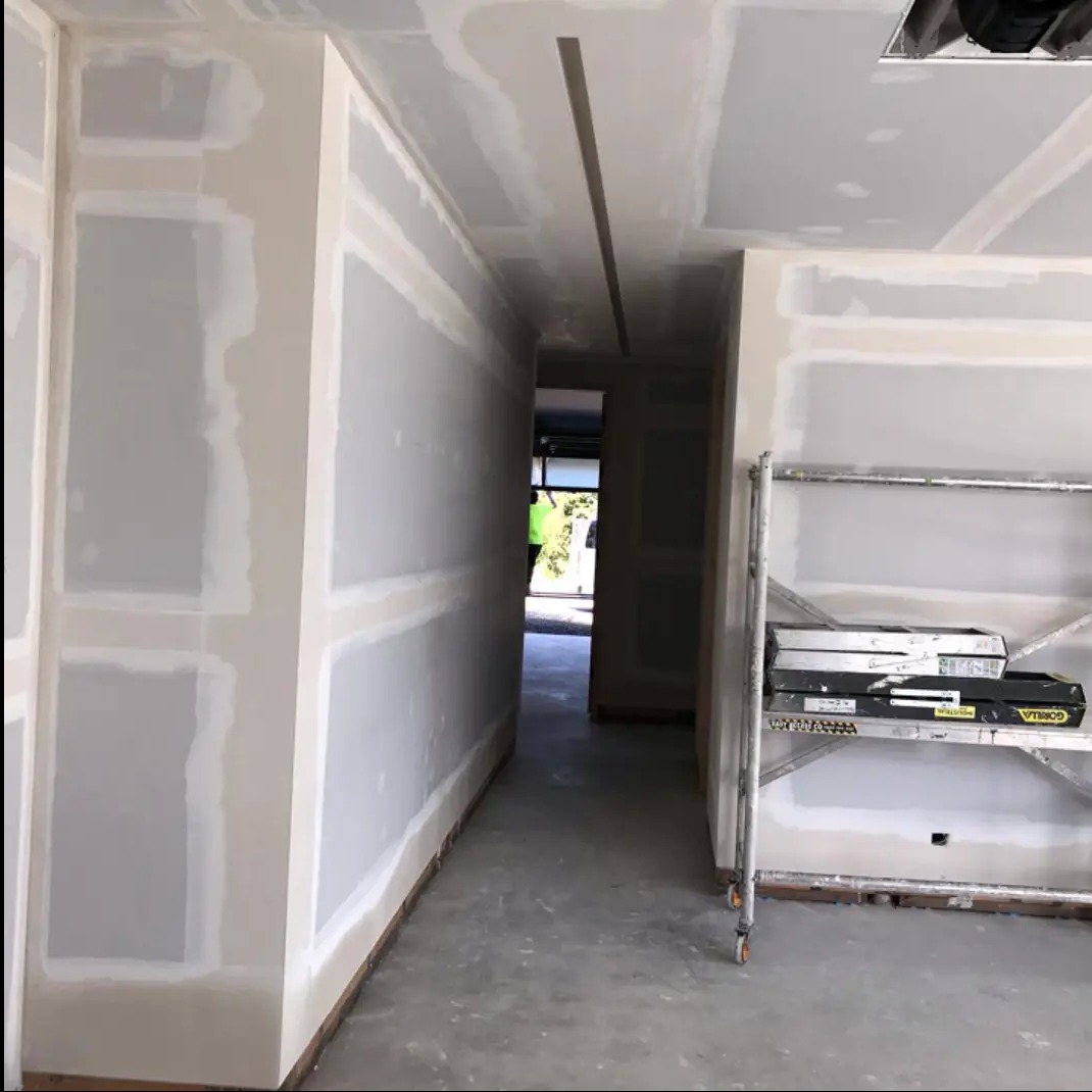 Korytarz w trakcie prac wykończeniowych: ściany z płyt gipsowo-kartonowych przygotowane do dalszych prac, widoczne łączenia i szpachlowanie, z boku rusztowanie z narzędziami.