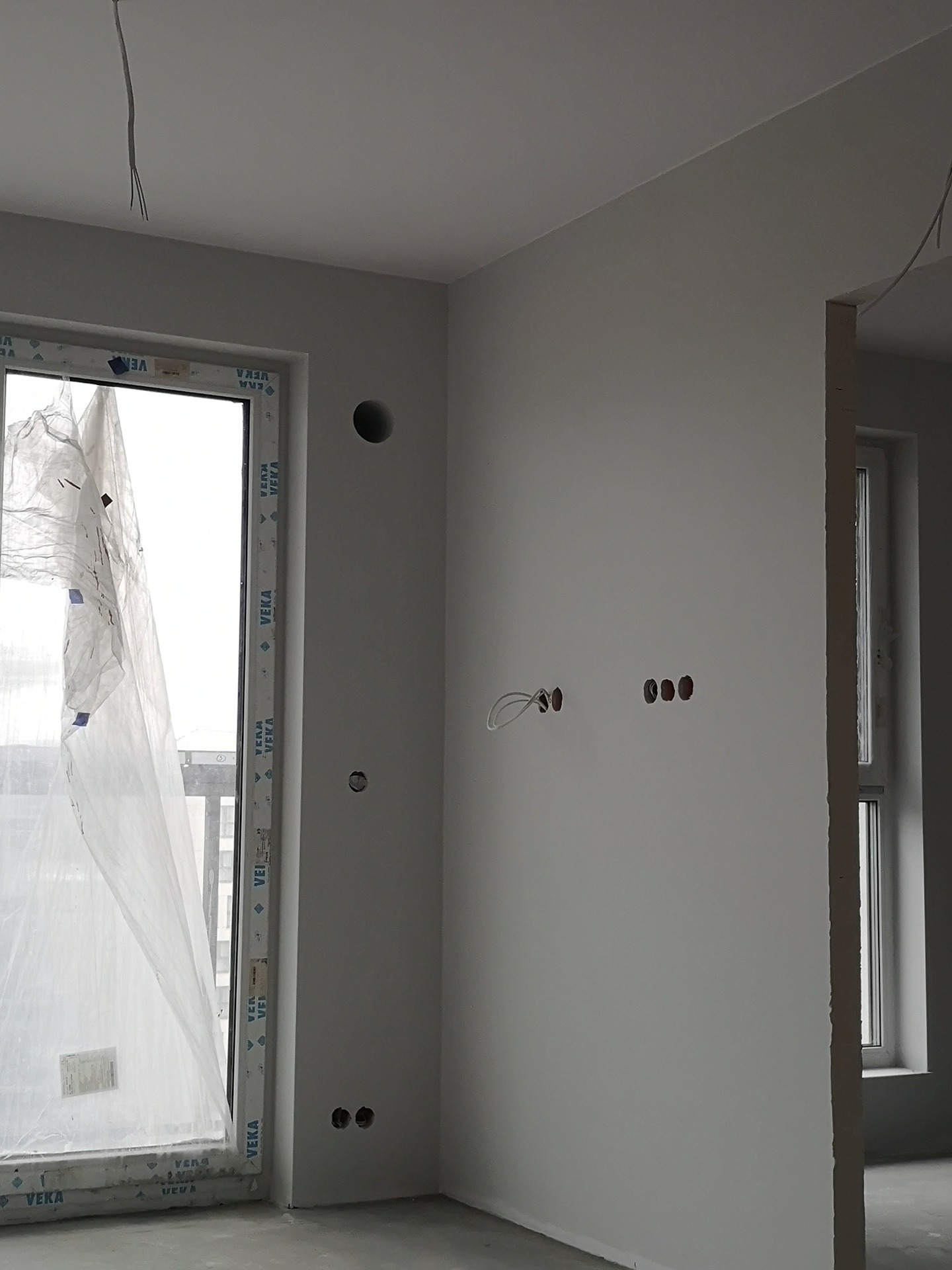 Świeżo pomalowane na szaro ściany w nowym mieszkaniu, widoczne otwory pod gniazdka elektryczne i osłonięte folią okno firmy VEKA.