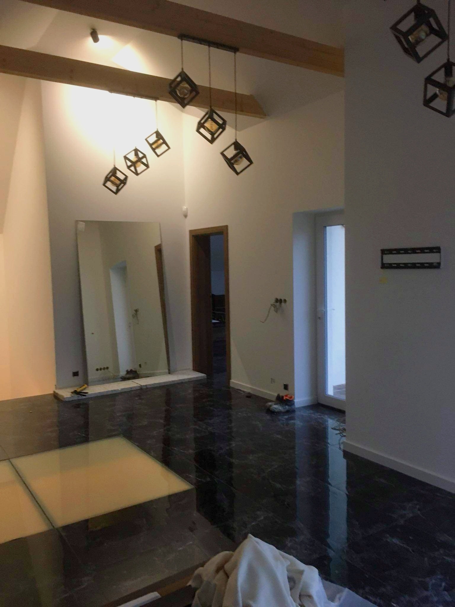Wnętrze w trakcie prac wykończeniowych: duże lustro oparte o ścianę, marmurowa podłoga, wiszące lampy w kształcie sześcianów, widoczne kable elektryczne na ścianie, fragmenty materiałów budowlanych...