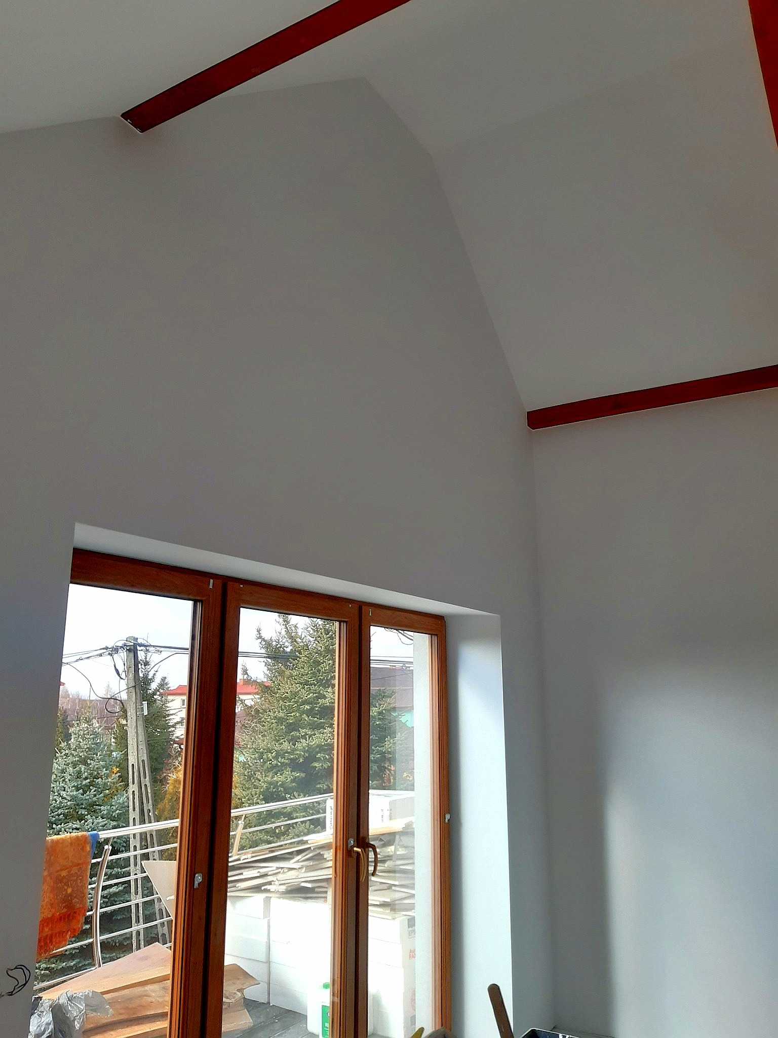 Pomalowane na biało poddasze z drewnianymi belkami na suficie i brązowymi oknami balkonowymi z widokiem na zewnątrz; częściowo widoczne narzędzia malarskie w dolnej części kadru.