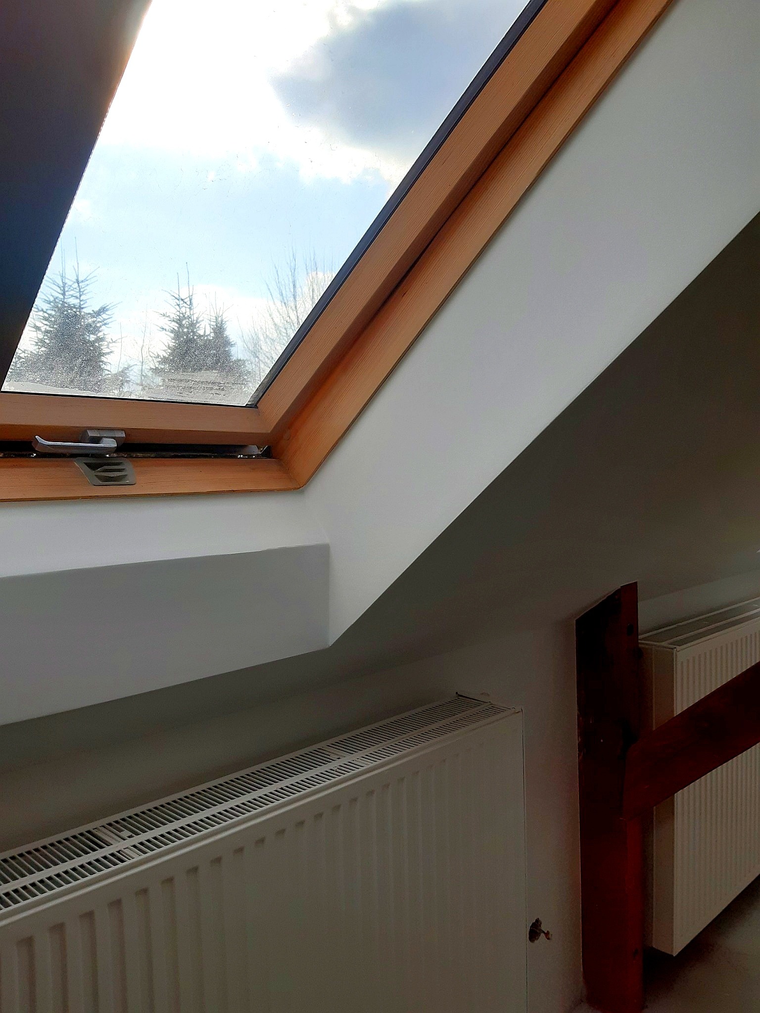 Poddasze z oknem dachowym z drewnianą ramą, widok na zewnątrz na drzewa, białe ściany, grzejnik pod oknem, drewniany element konstrukcyjny.