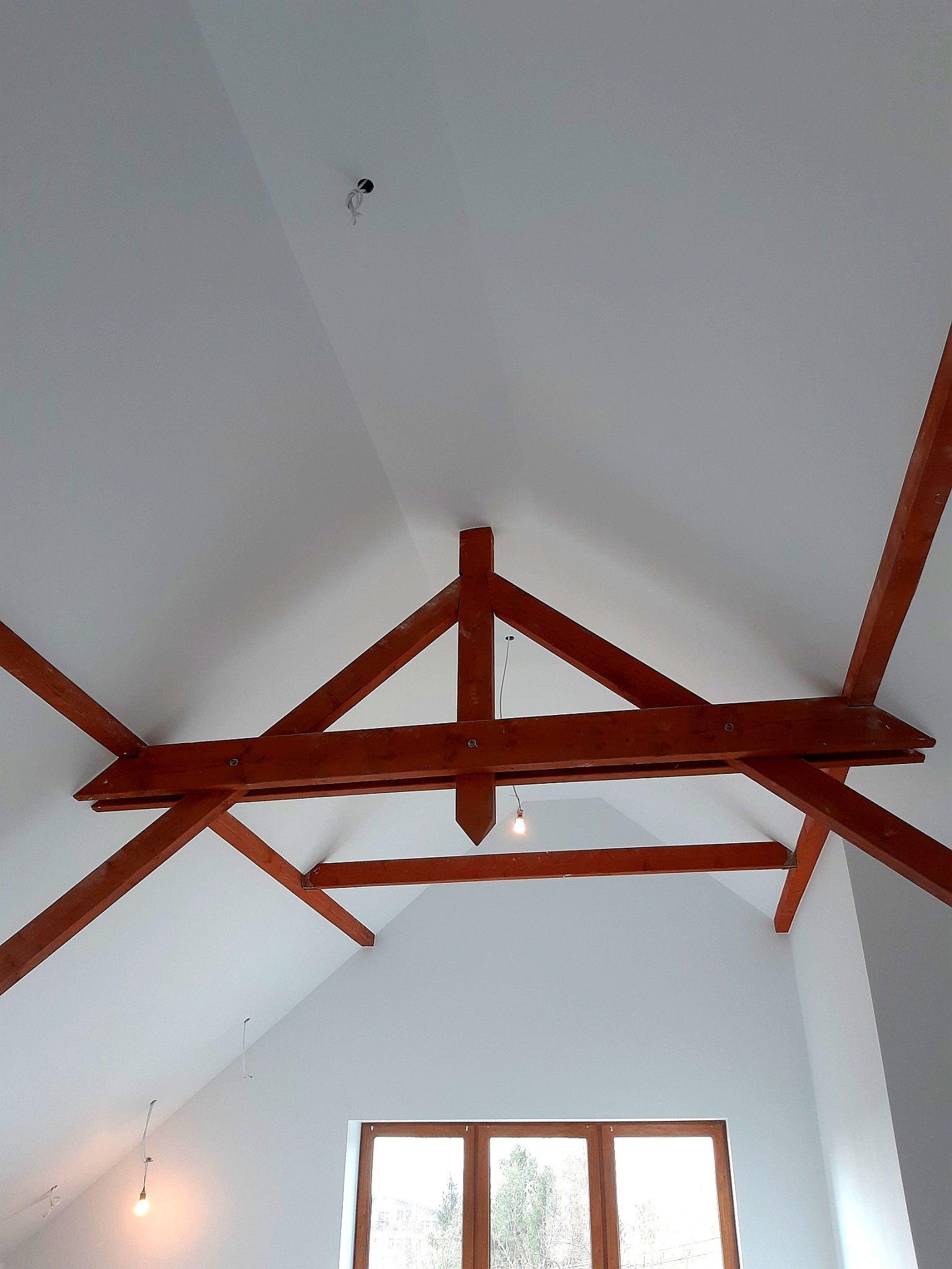 Wnętrze poddasza z drewnianą konstrukcją więźby dachowej, świeżo pomalowanymi na biało ścianami i sufitem, widoczne wyprowadzenia kabli elektrycznych i okno.